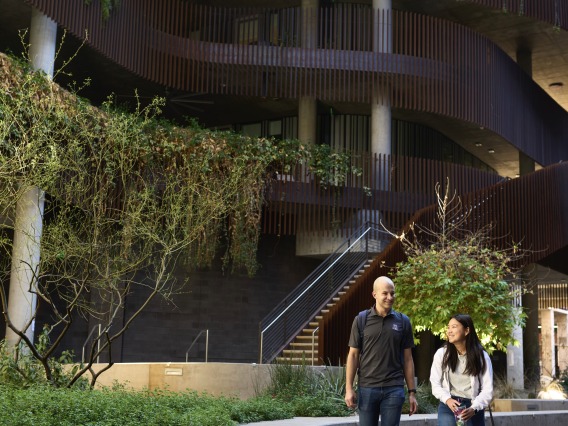 Two students walking on a pathway outside a building during the day. The pathway is covered in greenery