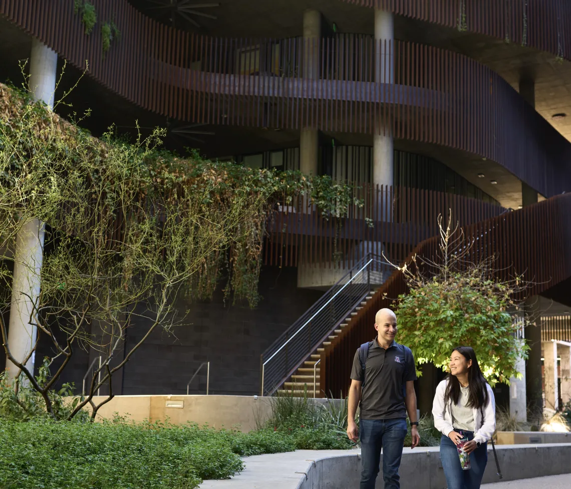 Two students walking on a pathway outside a building during the day. The pathway is covered in greenery